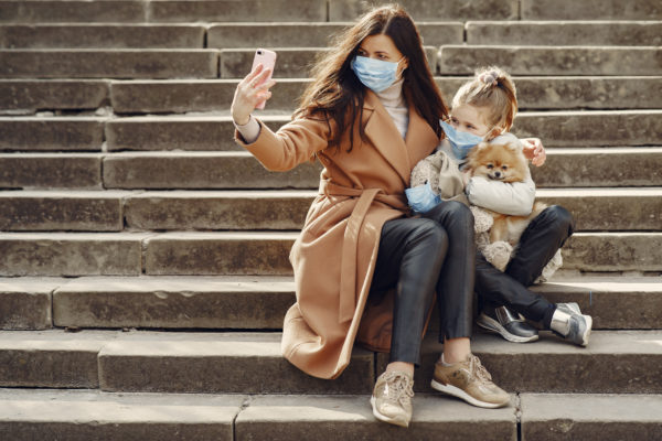 Canva - Young woman hugging cute daughter with dog in masks taking selfie on steps